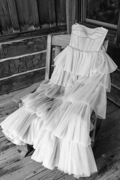 Wedding dress with tiered tulle skirt draped on a wooden chair, shown on a rustic porch with wood siding, window, and stone wall