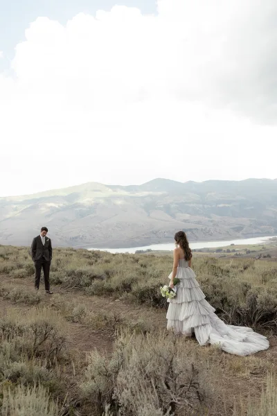 Couple portrait of bride and groom outdoors, she holds a green and white bouquet in a tiered dress before mountains and lake under cloudy sky