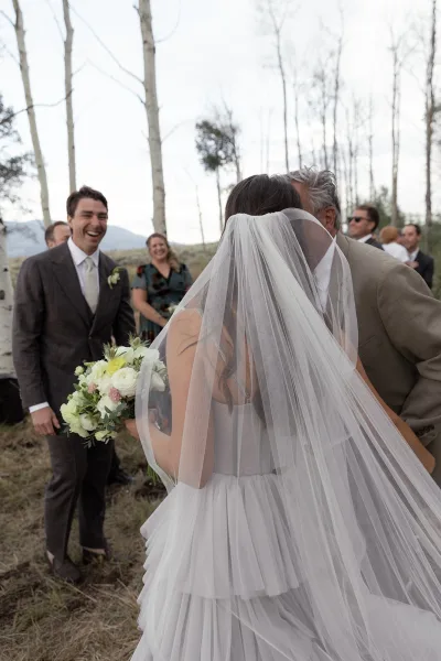 Wedding ceremony moment as bride greets her father, holding a bouquet with veil trailing, while guests watch in a mountain field under gray sky