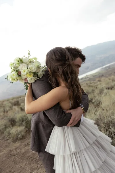 Wedding couple portrait of bride and groom hug, her white and green bouquet over shoulder, embracing on a mountainside above a lake