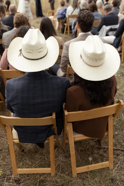 Wedding guests in western wedding attire seated on wooden folding chairs, some wearing white cowboy hats, at an outdoor ceremony on grass