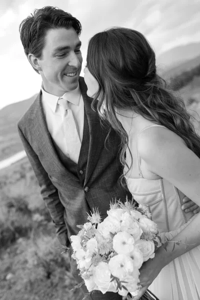 Couple portrait in black and white wedding portrait style, bride and groom leaning in to kiss holding bouquet against mountain hillside and sky backdrop