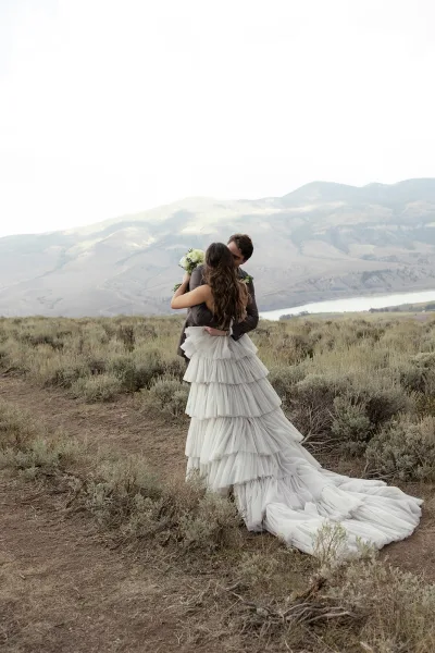 Wedding kiss as bride in strapless gown with long train holds bouquet, embracing groom in charcoal suit by lake and mountains under overcast sky