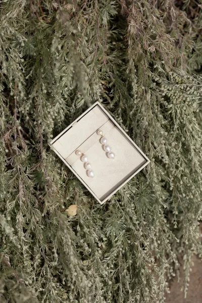 Wedding earrings with pearl drop earrings resting in a jewelry box, styled beside soft greenery foliage for a garden-inspired detail flat lay