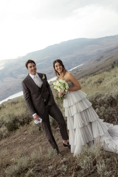 Couple portrait of newlyweds smiling, bride holding a white and green bouquet beside groom in brown suit with mountains and river behind
