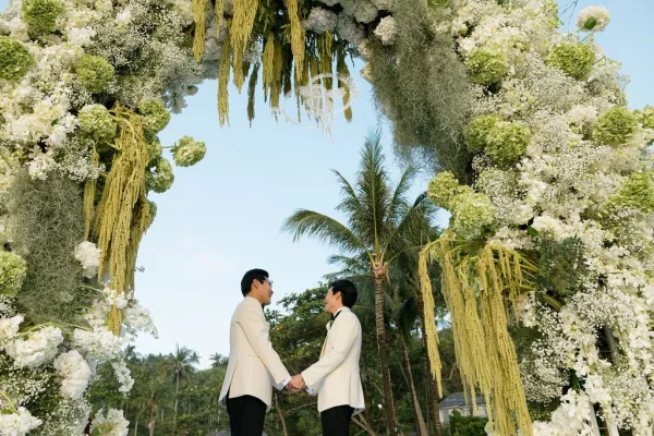 Ceremony moment with grooms holding hands under a floral arch, palm trees and blue sky behind them in an open-air garden setting.