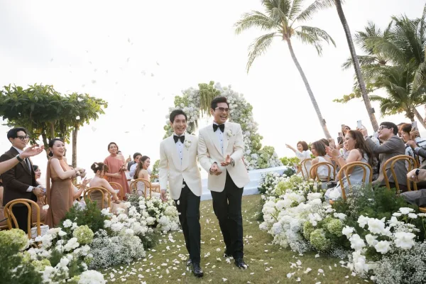 Wedding recessional during a joyful ceremony exit as guests toss flower petals, with palm trees lining the lawn beneath a bright sky.