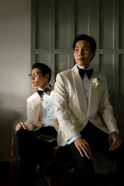Couple portrait of two grooms in tuxedos, seated close with calm expressions, under dramatic lighting against a paneled indoor wall.