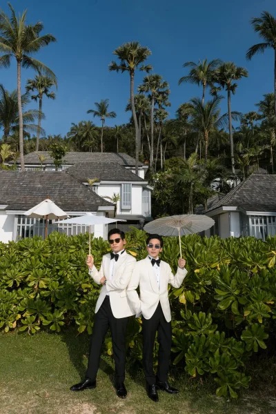 Couple portrait of two grooms, one holding a parasol, by a tropical villa framed with palm trees, green hedges, and bright blue sky.