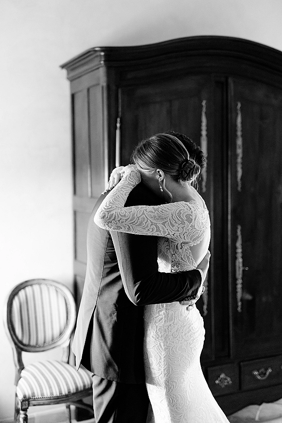 First look moment as bride and groom hugging, bride wiping tears in a lace long sleeve wedding dress beside a vintage armoire indoors