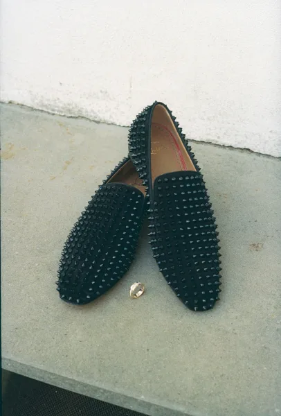 Groom shoes, black groom loafers with studded spikes beside a gold wedding band on a concrete ledge against a white wall