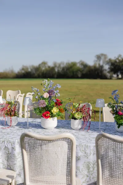 Reception tablescape with an outdoor reception table set in an open field, wildflower centerpieces in white ceramic vases and cane back chairs