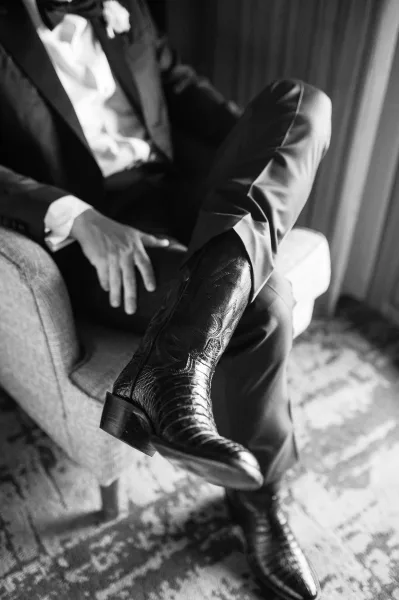 Groom details in a black tuxedo groom look, showing cowboy boots and boutonniere while seated by an armchair and curtain