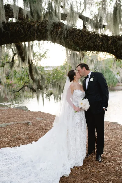 Wedding kiss portrait of bride and groom kissing under a moss-draped oak by a lake, with strapless lace dress, long veil, and white bouquet