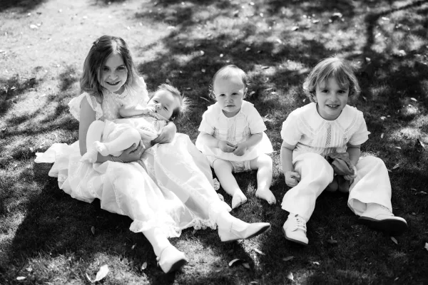 Flower girl portrait of a little girl in a white lace dress holding a baby on the grass lawn in tree shade with scattered leaves