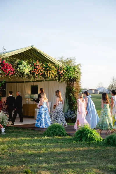 Wedding processional with bridesmaids walking down aisle on a wooden platform beneath a floral canopy in a sunny open field under blue sky