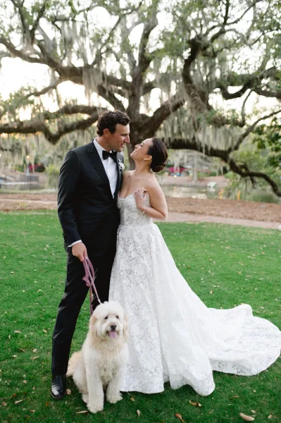 Couple portrait of bride in a strapless lace wedding dress and groom in tuxedo holding hands with a dog on a mossy oak lawn