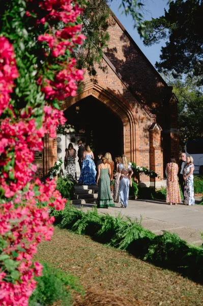 Wedding guests arriving at a brick church wedding entrance, walking past floral urns and greenery garland under the arched doorway in daylight