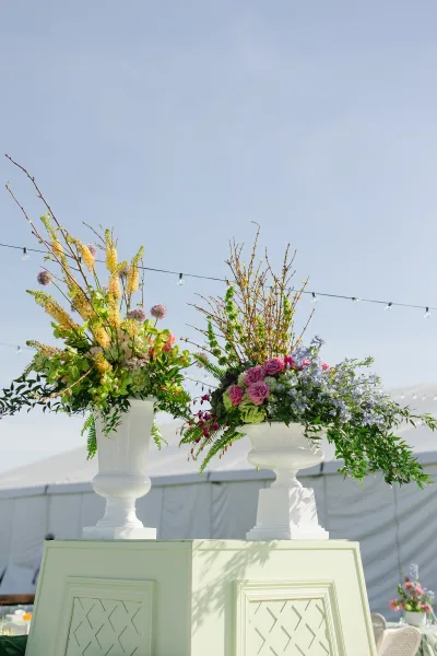 Wedding floral arrangements in white pedestal urns with greenery and branches, set beneath a tent canopy with string lights outdoors