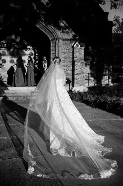 Bridal portrait of a bride walking up church steps, long veil trailing behind her as bridesmaids wait beneath a floral arch doorway