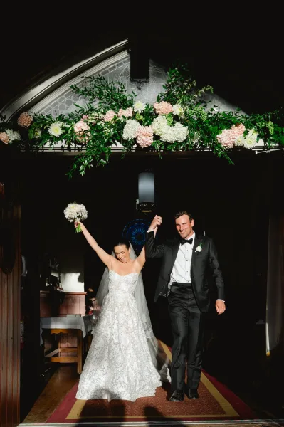 Wedding recessional as bride and groom hold hands, bride lifting bouquet beneath a floral garland in a stained glass hallway doorway