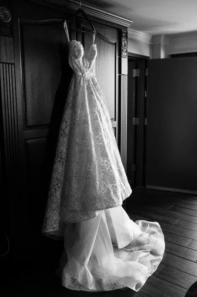 Wedding dress with lace detailing and spaghetti straps hanging on a hanger against a wood-paneled door in an interior hallway