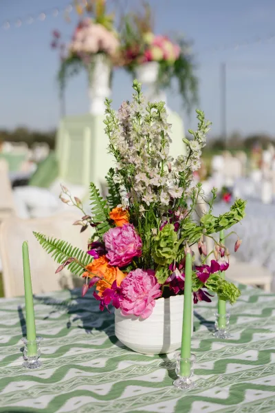 Wedding centerpiece with peonies and roses in a ceramic vase, paired with green taper candles on a patterned tablecloth outdoors