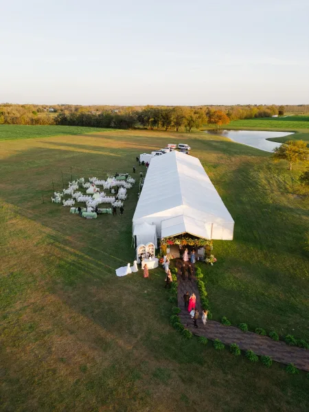Outdoor wedding reception under a white event tent with string lights, round tables, and wood dance floor beside a pond at sunset