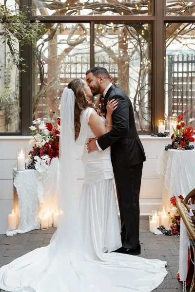 Wedding kiss as bride in long veil and satin gown embraces groom in black tuxedo, candlelit with string lights by greenhouse windows