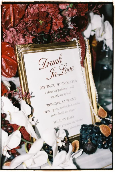 Wedding bar menu and signature cocktail menu in a gold frame with red flowers, white orchids, pearls, figs and grapes on a dark tabletop