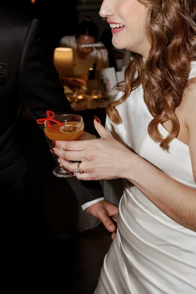 Bride cocktail moment at wedding cocktail hour, holding a coupe with red ribbon garnish beside candlelit reception tables and guests
