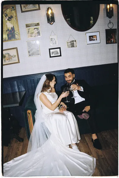 Wedding couple toast with champagne glasses, bride in satin dress and veil and groom in tux on lounge couch beneath wall sconces