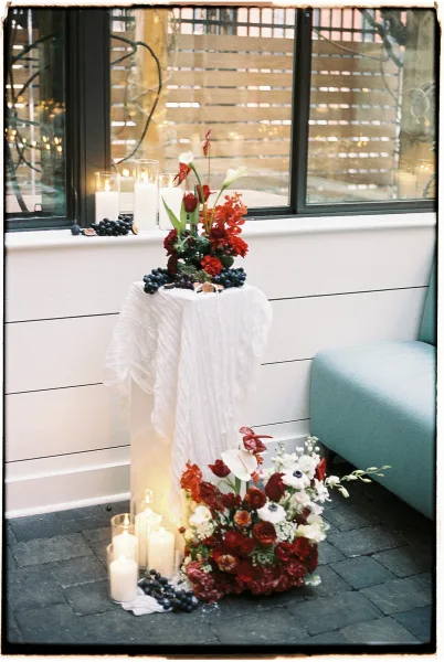 Wedding floral arrangement with red and white blooms and greenery, paired with pillar candles in glass cylinders by a window with string lights