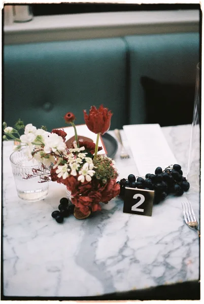 Reception tablescape with wedding table centerpiece of red tulips and white blooms, grapes, menu card, and black table number on marble table