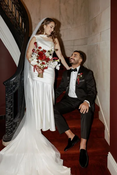 Couple portrait of bride and groom on stairs, bride in one-shoulder gown with veil, holding cascading bouquet on red-carpeted staircase