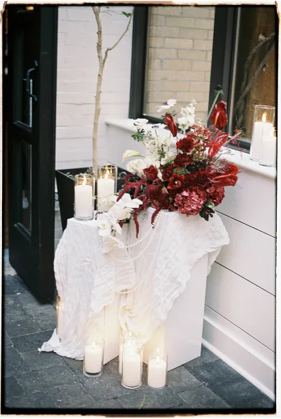 Wedding floral arrangement with red and white wedding flowers atop a pedestal, flanked by glass pillar candles and draped fabric by a brick wall