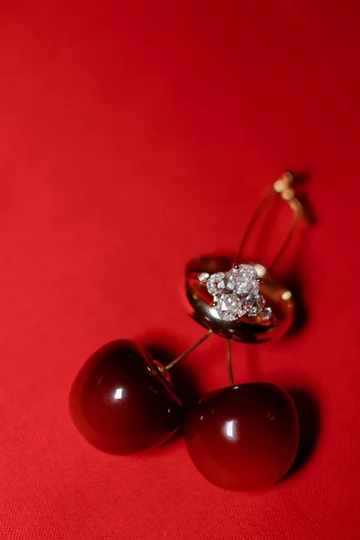 Engagement ring with oval diamond set on a gold band, styled on red fabric backdrop with cherry earrings for bold romantic detail