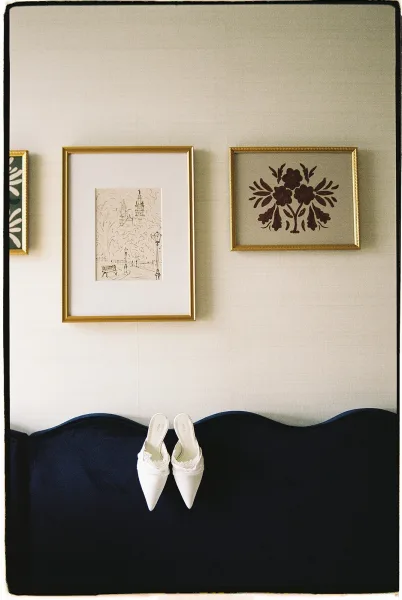 Bridal shoes, white wedding heels with lace trim, resting on a navy upholstered sofa beneath vintage framed wall art in a bright room