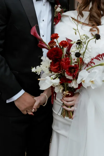 Wedding bouquet of red and white bridal bouquet blooms with calla lilies and orchids, held by bride and groom in black tie against dark backdrop