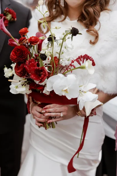 Bridal bouquet with red and white wedding bouquet blooms, white orchids and burgundy ribbon, held by bride in faux fur wrap on walkway