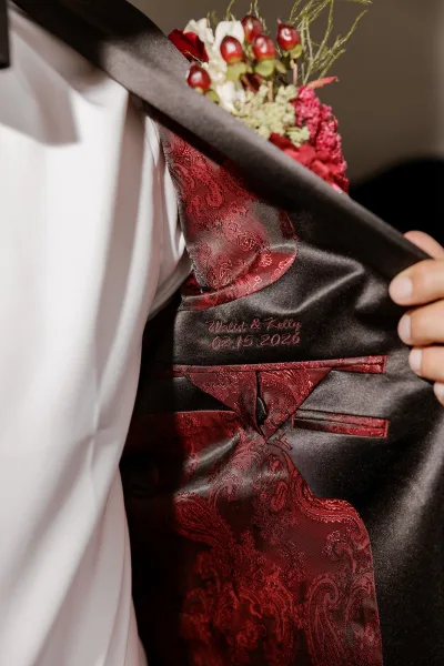 Groom suit details with embroidered wedding suit lining, showing black jacket, burgundy paisley vest and tie, boutonniere in indoor light