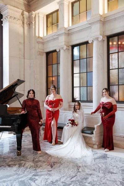 Bridal party portrait of bride with bridesmaids in red dresses, holding calla lilies by a grand piano in a marble hall