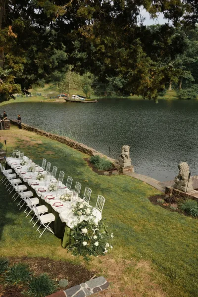 Outdoor reception tablescape with long banquet table decor, taper candles glowing along a grass lawn beside a serene lake and boats.