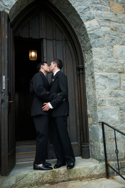 Wedding kiss portrait as two grooms kiss on stone steps beneath an arched wooden church doorway, in black tuxedos; one groom wears eyeglasses.