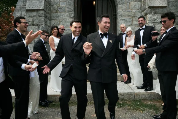 Ceremony exit during a wedding recessional, two grooms holding hands stride through confetti on stone church steps as guests cheer by wooden doors.