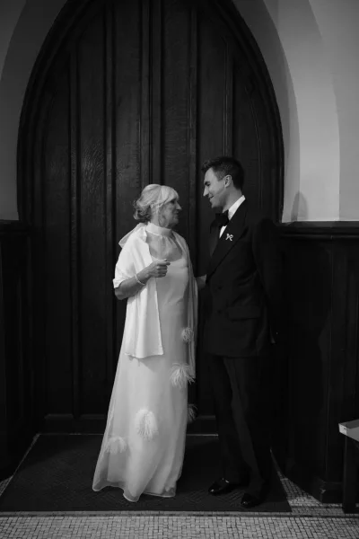 Wedding family moment as groom with mother share a candid embrace, his black bow tie crisp, framed by an arched wooden door and warm paneling.