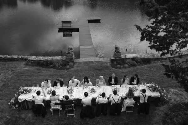 Reception tablescape on a long banquet table with lush florals, set on a lawn by a lake, with a stone wall, trees, and a dock behind.