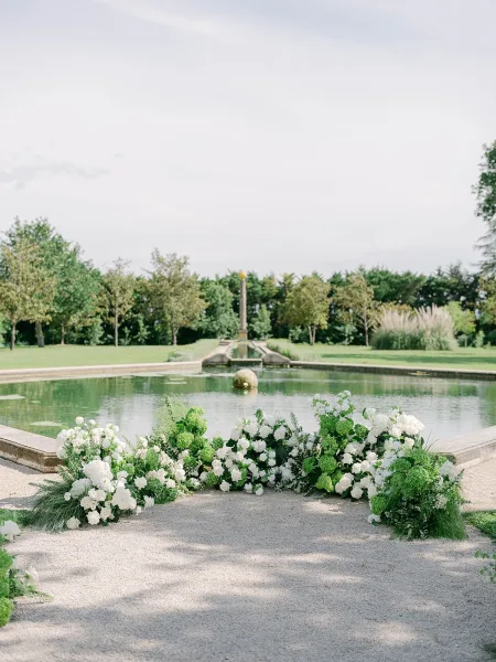 Ceremony floral installation of white florals framing a reflecting pool and fountain, refined outdoor ceremony decor amid garden trees.