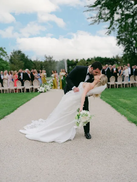 Wedding kiss portrait of a newlywed dip kiss on a garden lawn, bride clutching a white and green bridal bouquet with guests seated nearby.