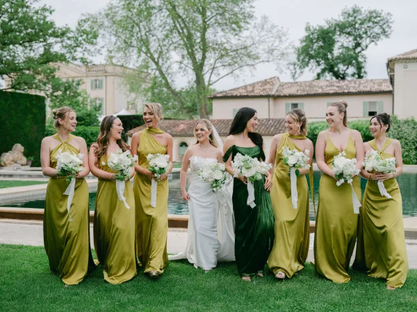 Bridal party portrait of a bride with bridesmaids, carrying white bouquets beside a reflecting pool and a tile-roofed villa garden.